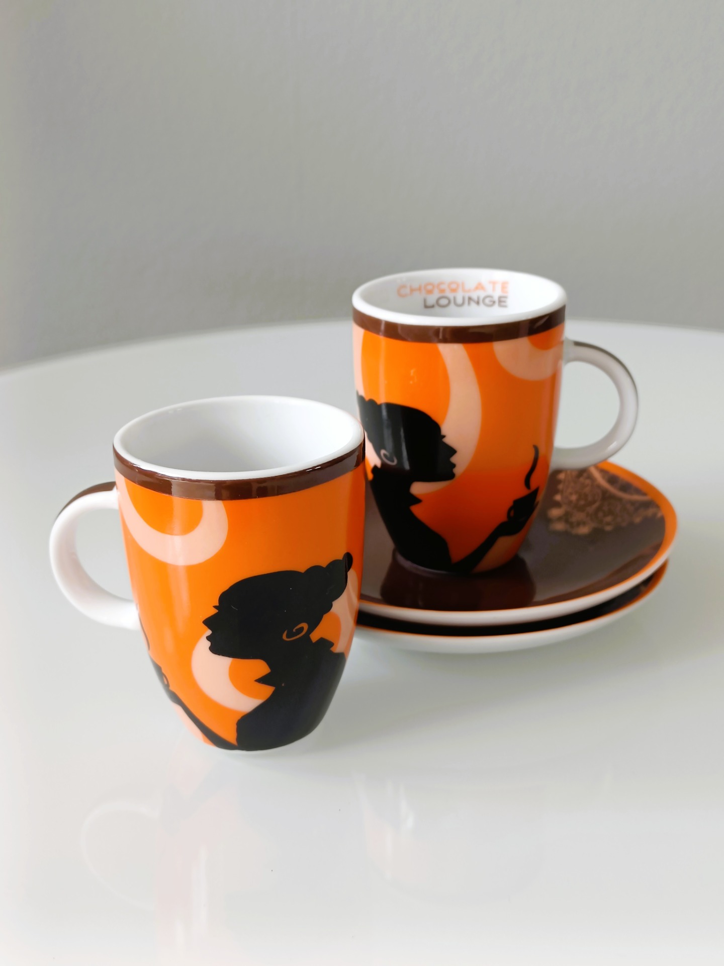 Twee oranje espressokopjes met zwarte vrouwenportretten en een schoteltje van het merk Chocolate Lounge, collector's item